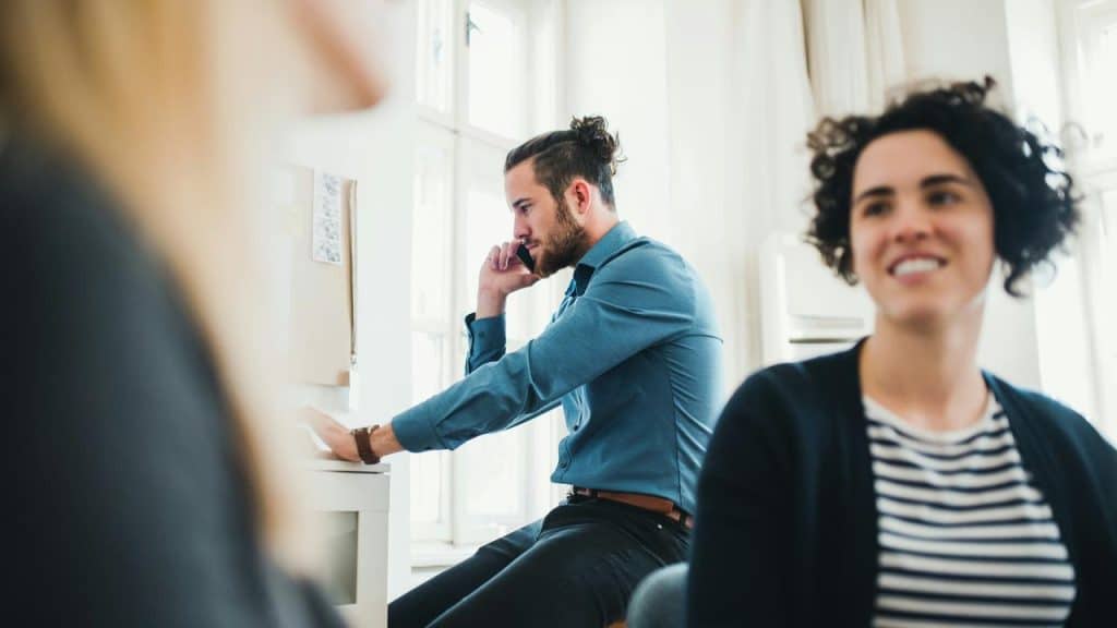 A man talks on the phone at his desk as two women sit nearby in an office space.