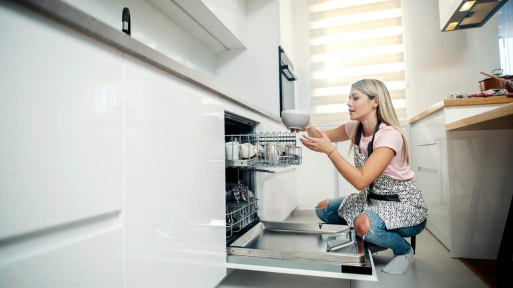 A woman wearing an apron unloads clean dishes from a modern dishwasher.