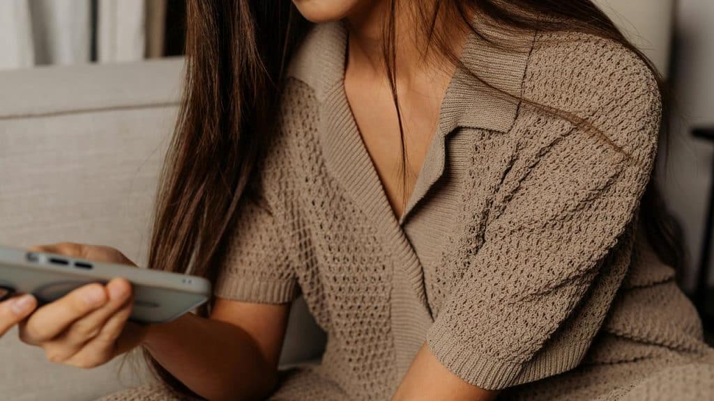 A woman sits on the couch in a knitted dress, holding her phone thoughtfully.