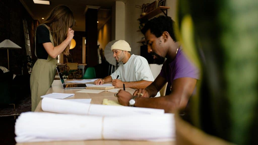 A group of three people work together at a table with notebooks and a laptop.