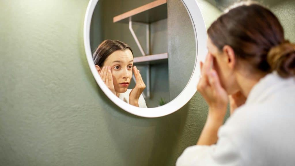 A woman looks at her reflection closely in the mirror, adjusting her face or hair.
