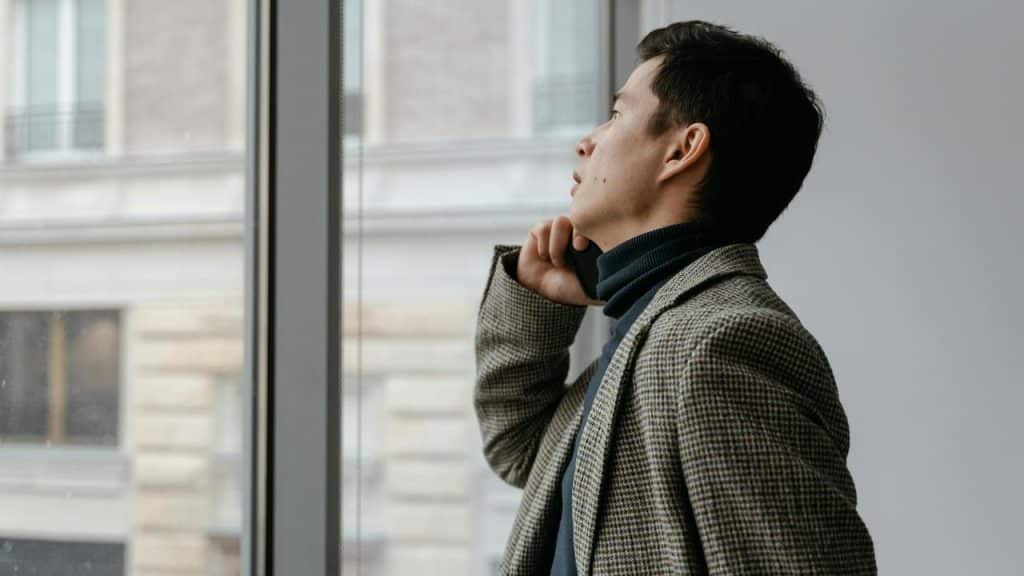 An adult man in a checked blazer talks on the phone while looking out a window.