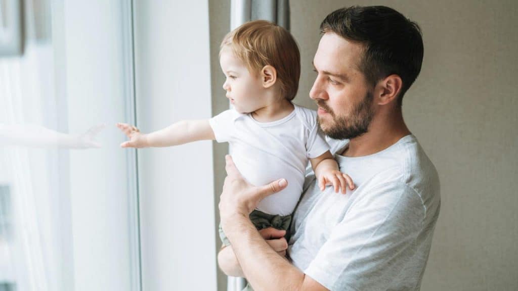 A father standing by the window and holding his child close.