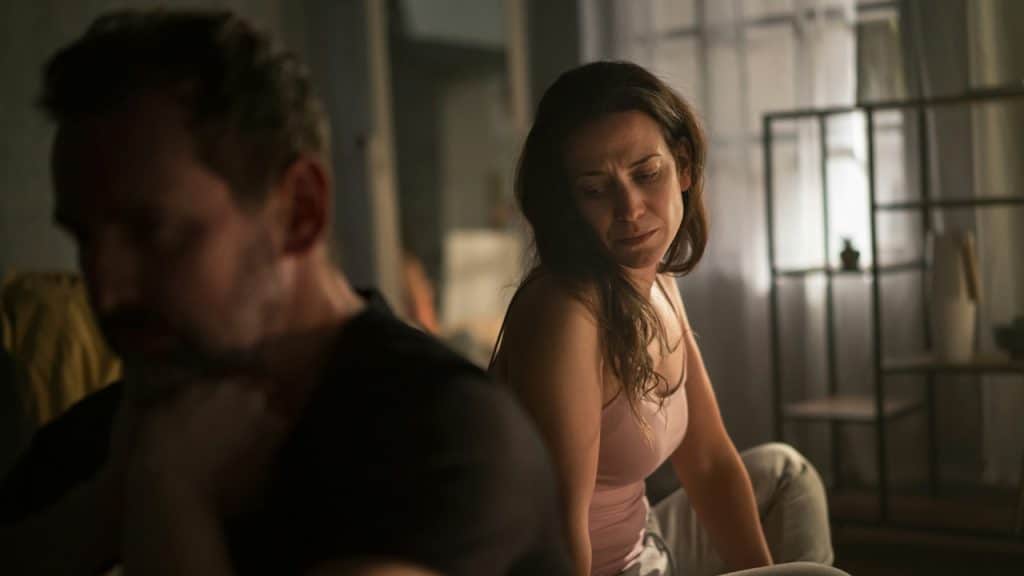 A woman looking distressed while sitting on a bed, with a man blurred in the foreground.