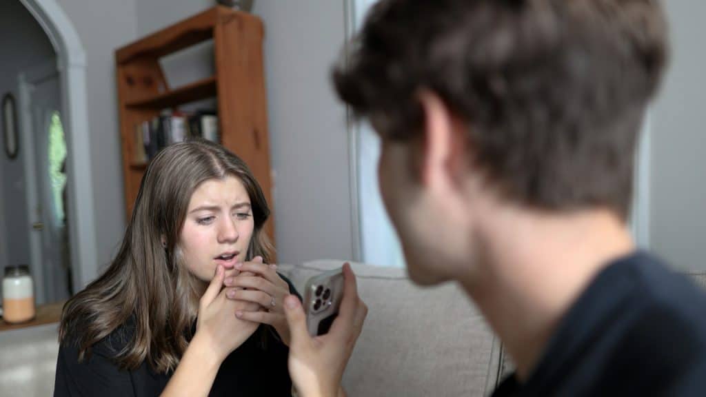 A woman sitting on a couch looking surprised or upset as a man, out of focus, shows her something on his phone.