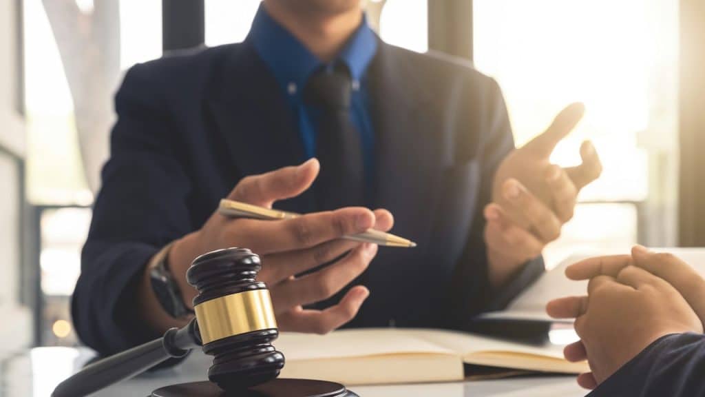 Close-up of a gavel on a white table, with a lawyer in a suit talking to a client.