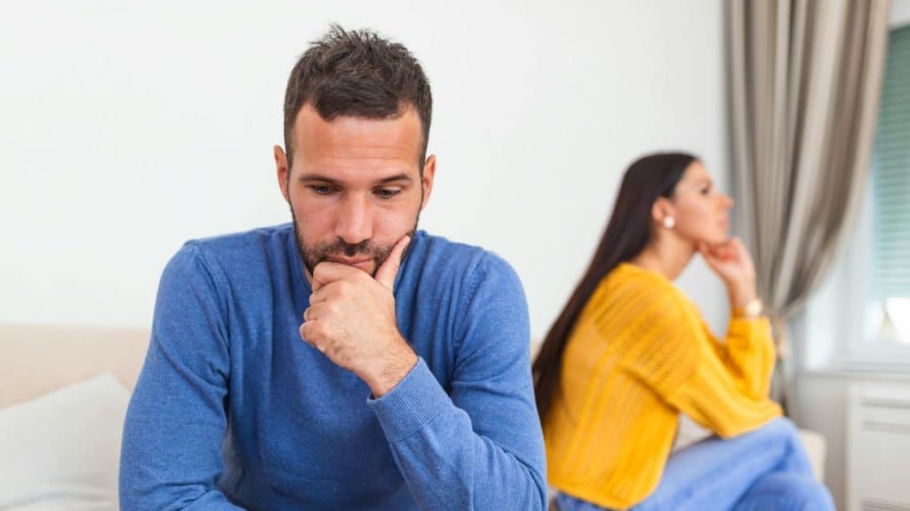 A man in a blue sweater sitting with his chin resting on his hand, looking troubled, with a woman in a yellow sweater blurred in the background.
