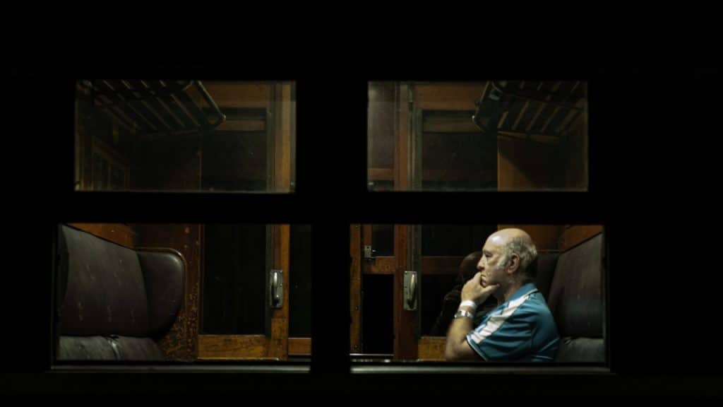 An elderly, bald man sitting alone in a dark train carriage, looking thoughtful with his hand on his chin.
