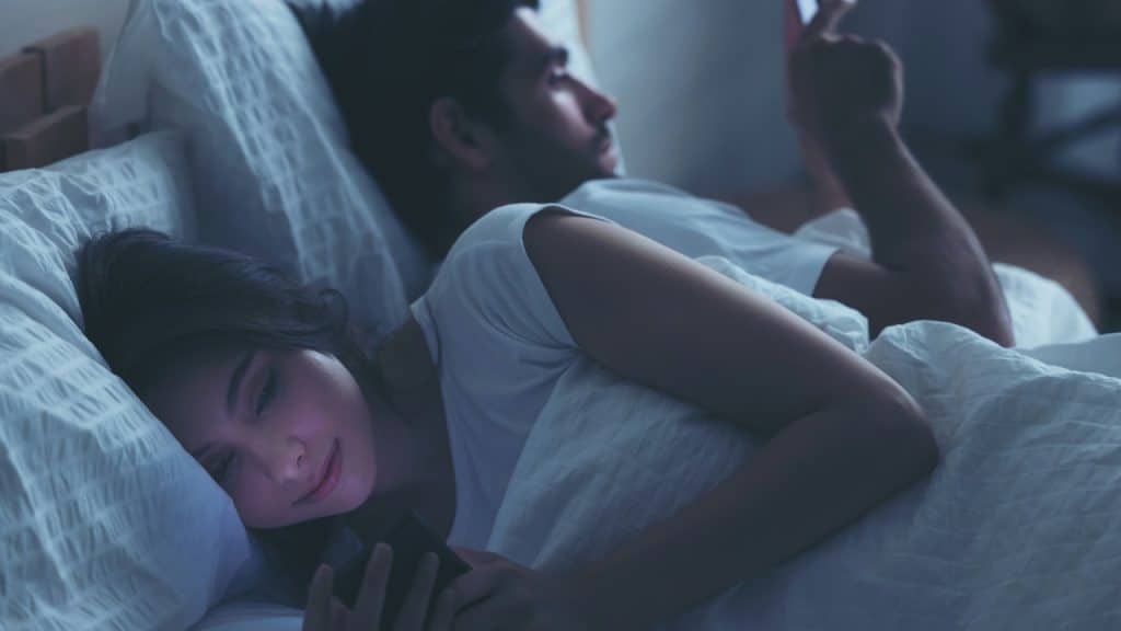 A couple lying in bed in a dark room, both looking at their phones.