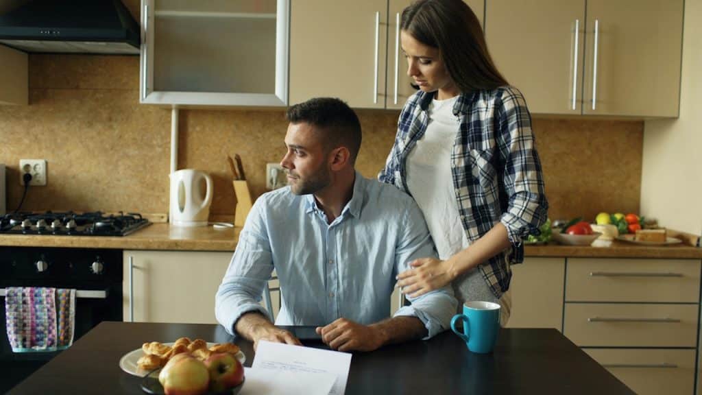A woman comforting or talking to a distressed man seated at a kitchen table with papers and snacks.