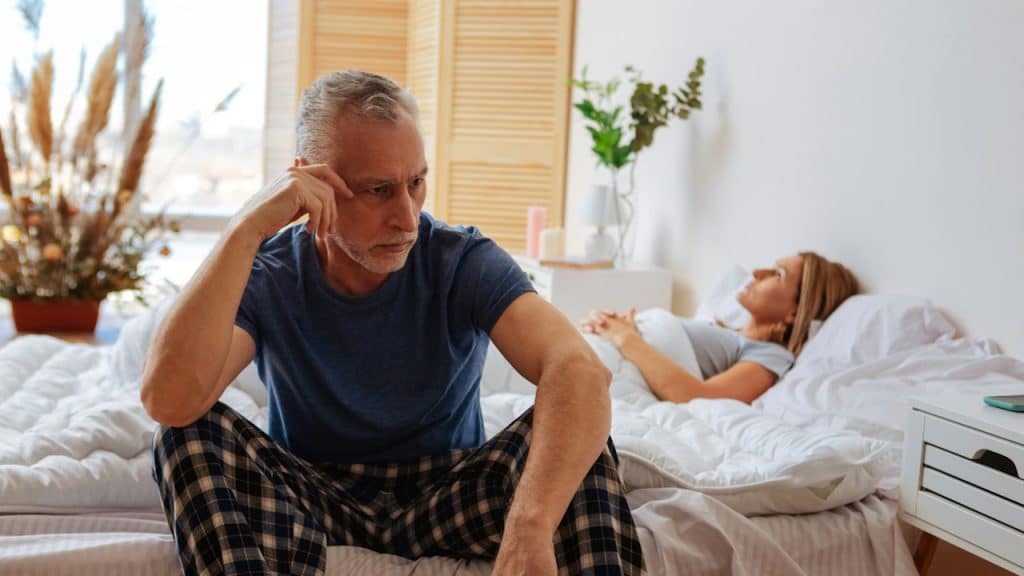 A distressed older man is sitting on the edge of a bed in plaid pajamas, while a woman lies asleep in the background.