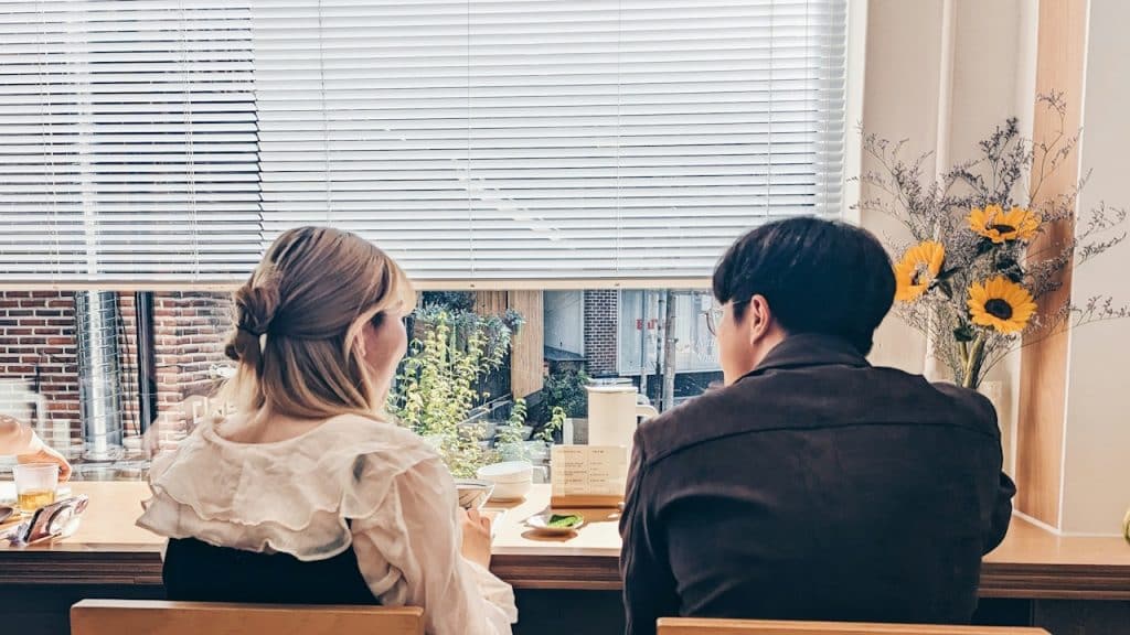 Two people sitting with their backs to the camera, looking out a window with blinds, next to a vase of sunflowers in a cafe.