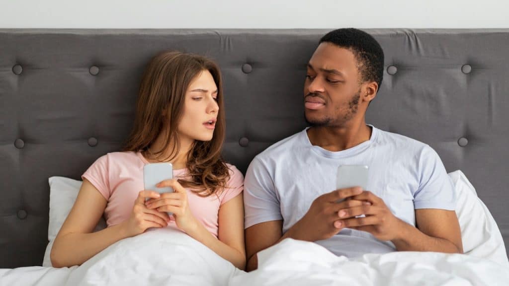 A man and a woman lying in bed, both holding smartphones, with the man looking suspiciously at the woman's screen.