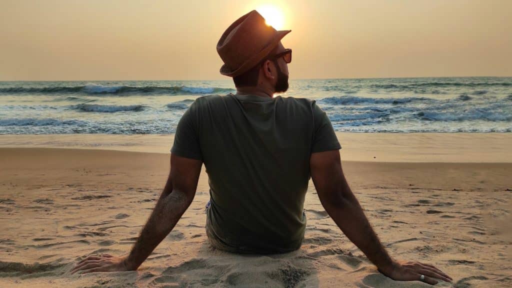 A man wearing a fedora and sunglasses sitting on the beach, facing the ocean sunset.