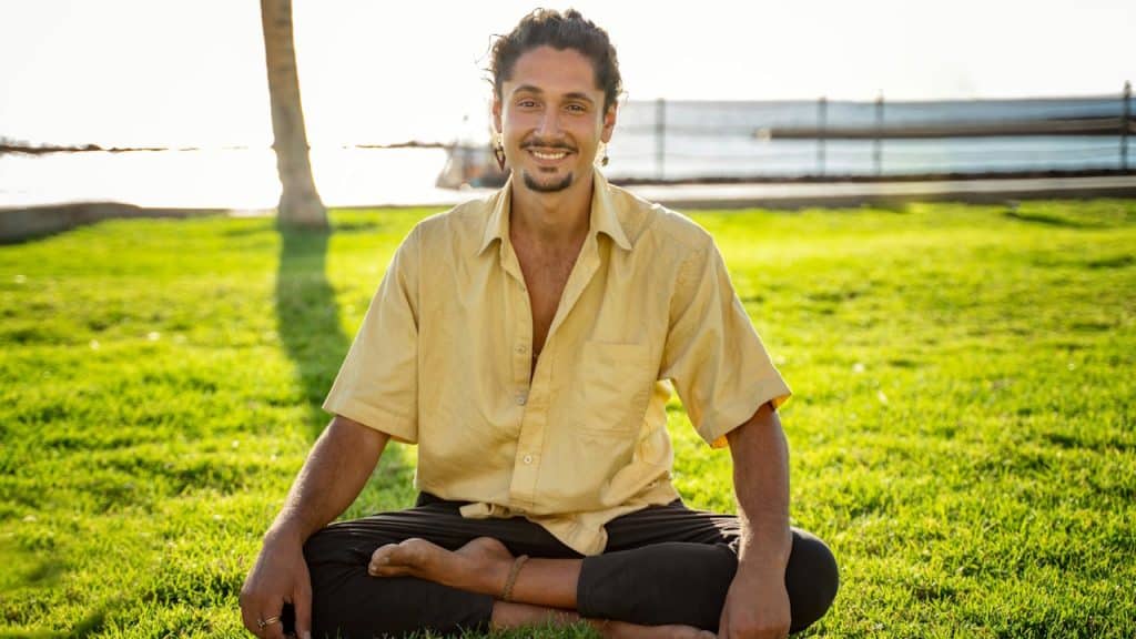 A smiling man with a beard and long hair sitting cross-legged on bright green grass near the ocean.