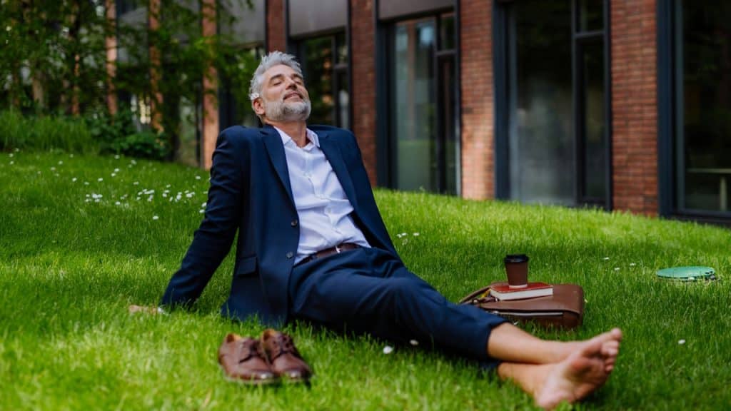 Barefoot senior businessman in a dark suit relaxing on grass outside an office building.