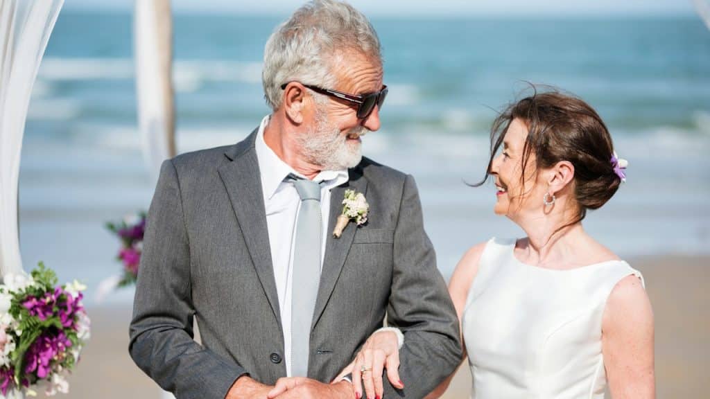 Older couple smiling at each other on a beach wedding day, with the man in a grey suit and the woman in a white dress.