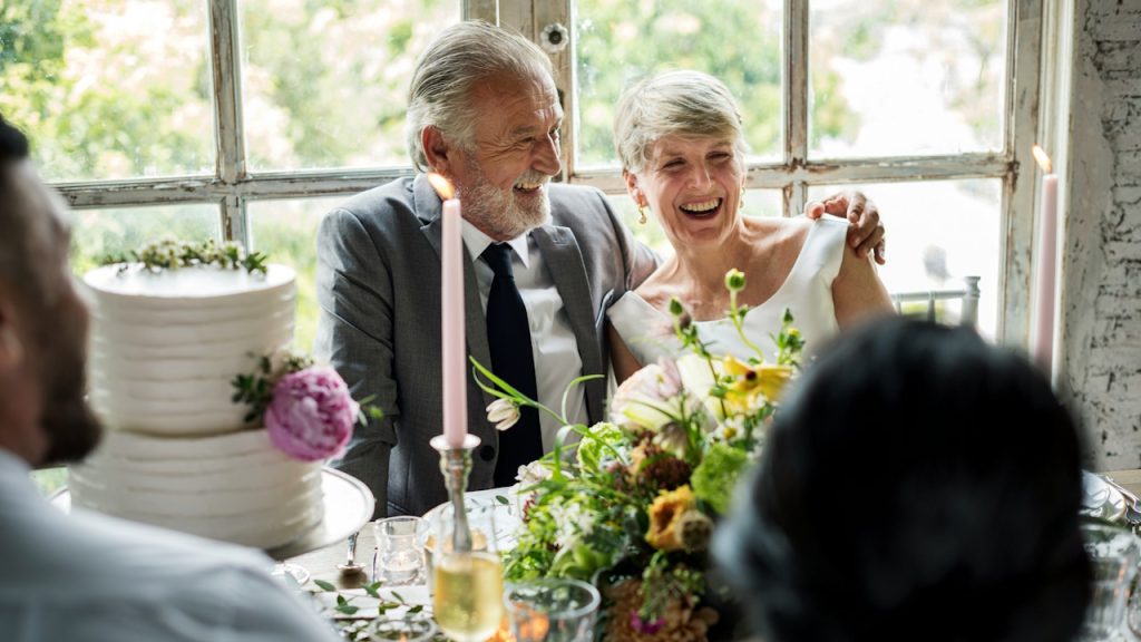 An elderly couple, dressed formally, smiling and laughing at aย wedding reception tableย with a tiered cake and flowers.