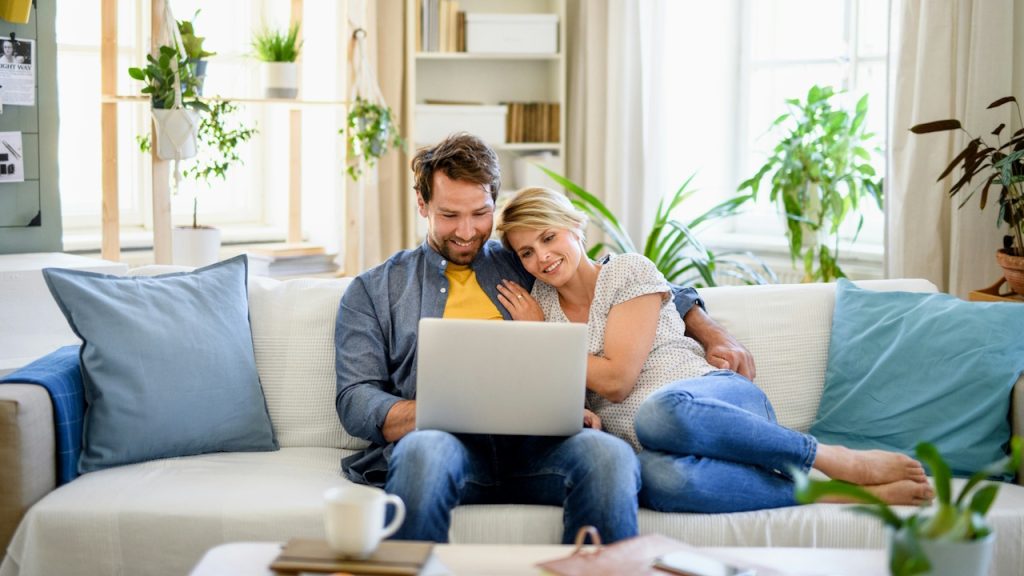 Happy couple embracing on a bright couch in a sunny living room, looking at a laptop together.