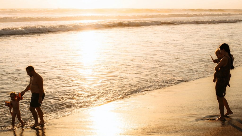 Family on a beach at sunset, with a man and young child walking towards the water and a woman holding another child.