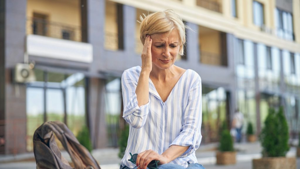 A middle-aged woman sitting outdoors with her hand pressed against her temple, looking distressed.