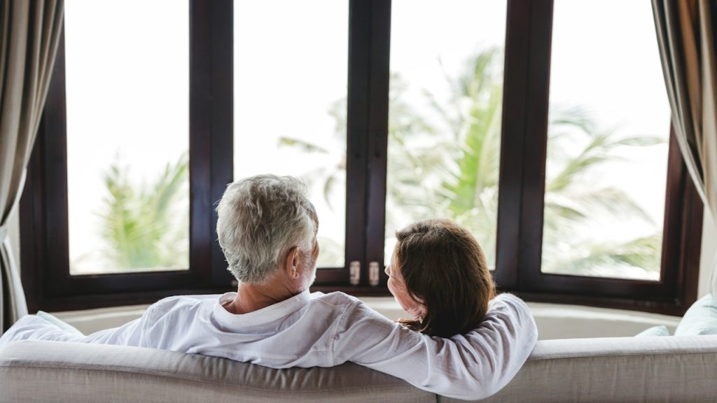 An older couple sitting on a couch, seen from behind, looking out a large window at palm trees and bright light.