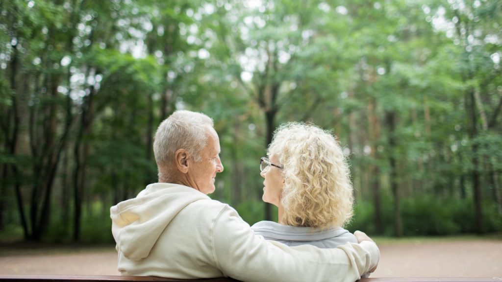An elderly couple sitting on a bench in a forest or park, facing each other and sharing a moment with the man's arm around the woman's shoulder.