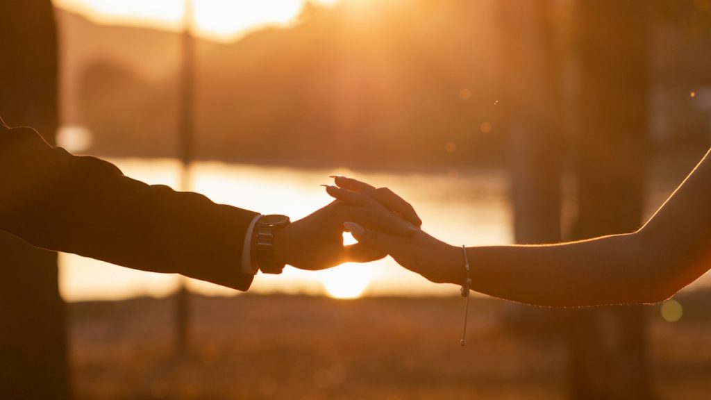 A close-up of a man's and a woman's hands gently touching or holding, silhouetted against a bright, golden sunset.