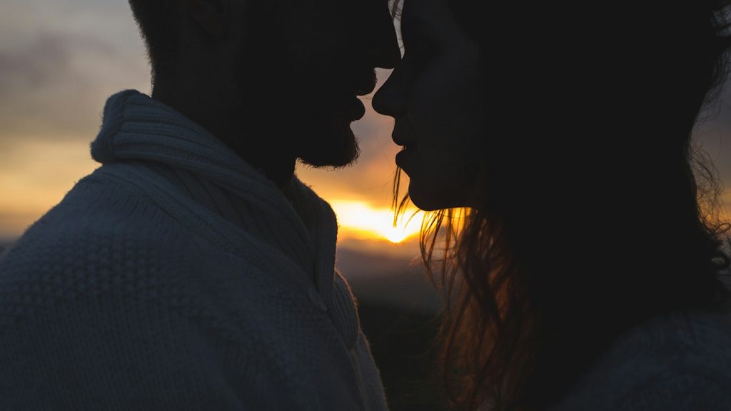 A romantic silhouette of a couple standing nose-to-nose, about to kiss, with the sunset glowing brightly between them.