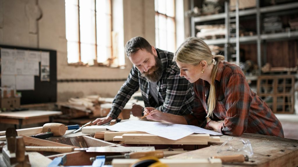 Two carpenters, a man and a woman, looking at blueprints indoors in a carpentry workshop.