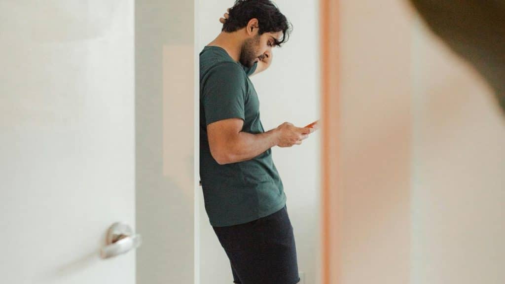 A man leaning against a wall looking down at his phone in deep thought.