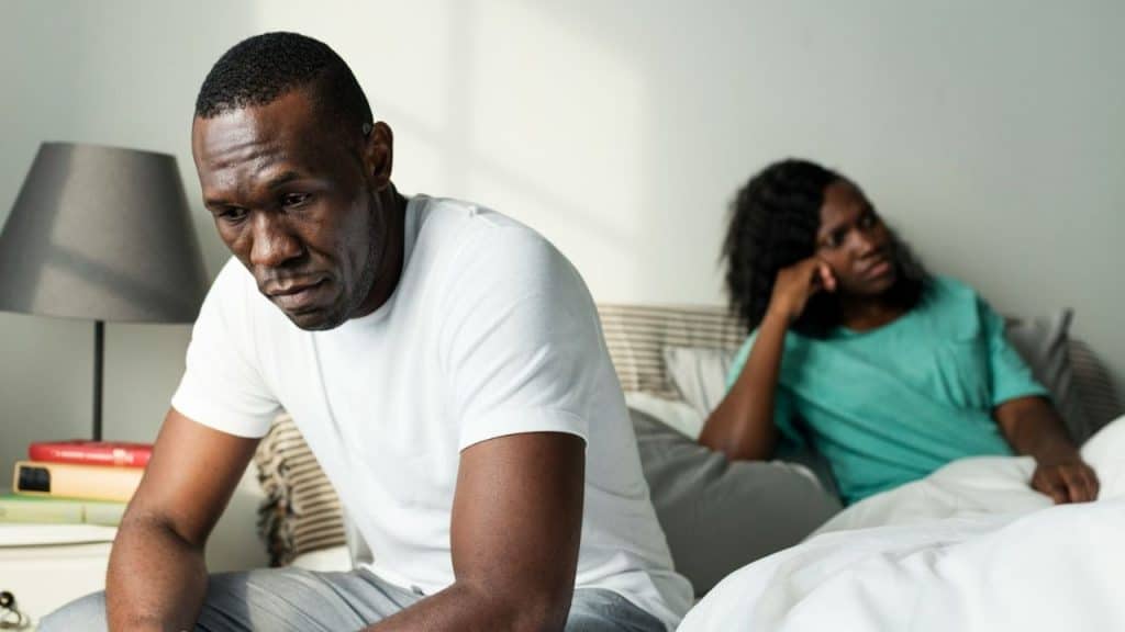 Close-up of a sad man sitting on a bed with a woman sulking in the background.