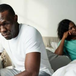 Close-up of a sad man sitting on a bed with a woman sulking in the background.