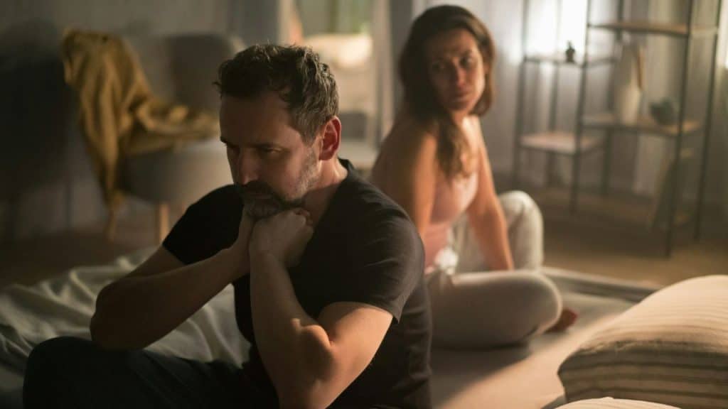 Man looking sad with chin on hands while sitting on floor, back to a woman.