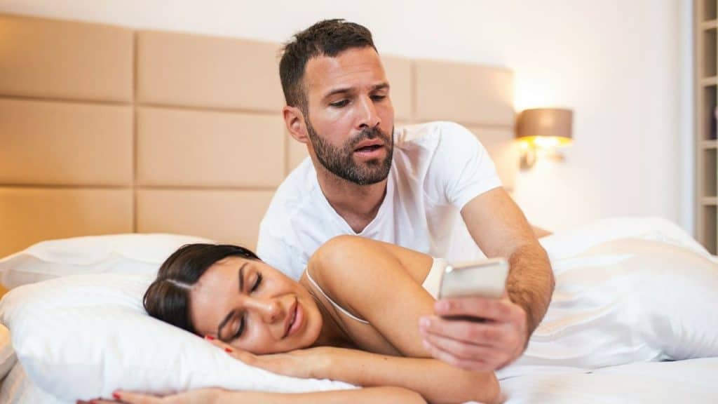 Man showing his phone screen to a woman lying down in bed, looking surprised.