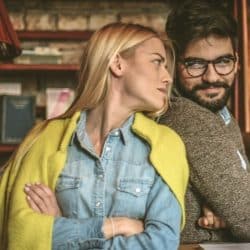 Blonde woman and bearded man with glasses looking at each other indoors.