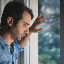 A pensive man in a denim shirt looks out a window with his hand on the glass.