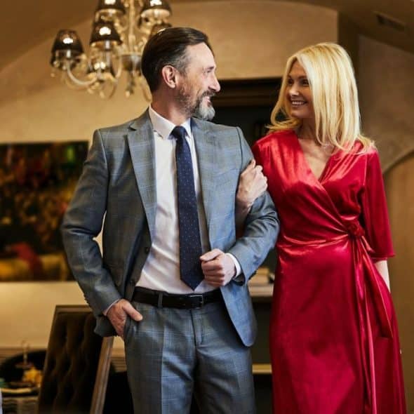 A formally dressed, middle-aged man and woman smile at each other indoors.