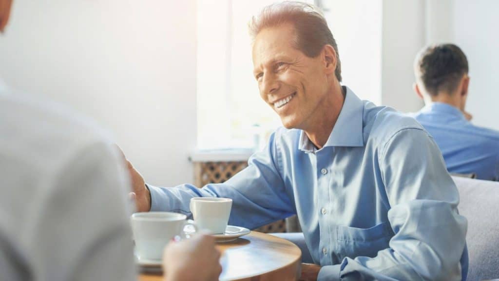 A handsome, middle-aged man in a blue shirt is smiling while talking over coffee.