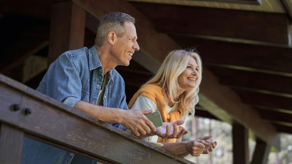 A middle-aged man and woman smile while leaning on a wooden balcony railing.