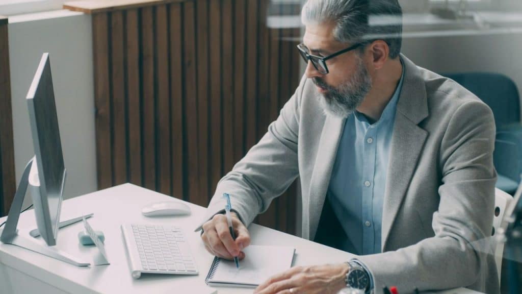 A middle-aged, bearded man in a blazer writes in a notebook at a desk.
