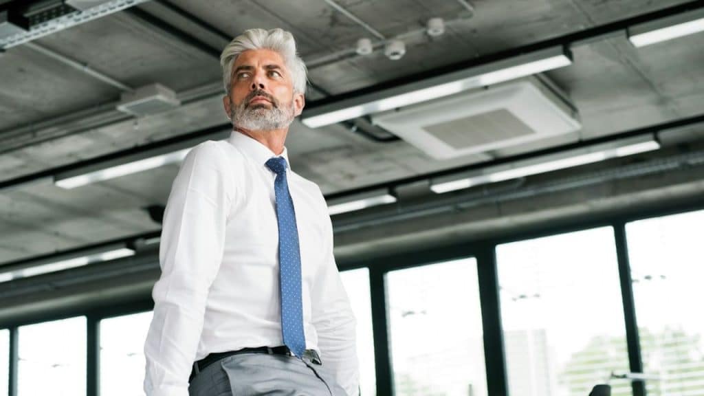 A well-dressed, silver-haired man looks away in a modern office setting.