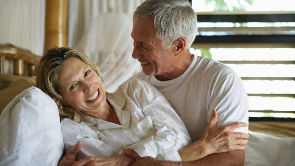 A happy older couple smiles and embraces while sitting up in bed.