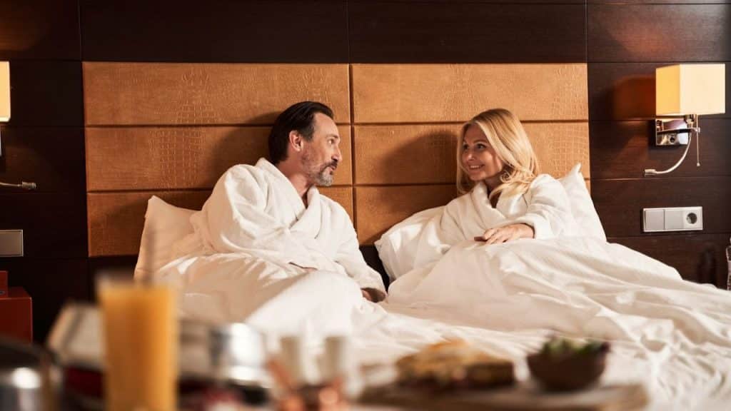 A smiling middle-aged couple in bathrobes talks while sitting up in a hotel bed.