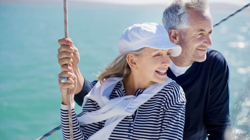A smiling, middle-aged woman in a striped shirt and cap holds a rope on a sailboat next to a man.