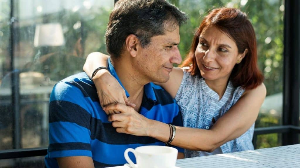 A smiling woman embraces a man from behind as they sit at a table.