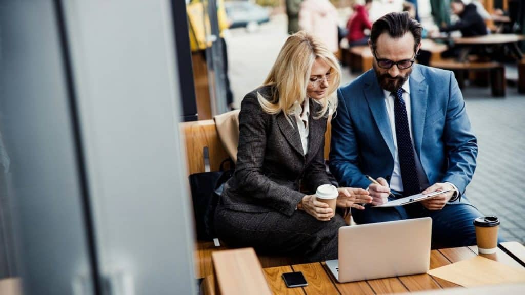 A professionally dressed man and woman review documents at an outdoor table with a laptop.