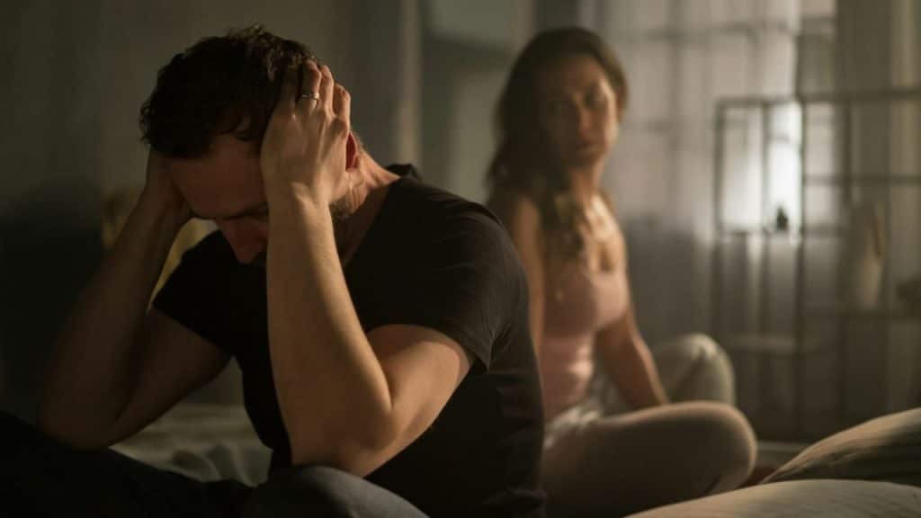 A man sits with his head in his hands, looking down, while a woman sits blurred in the background.