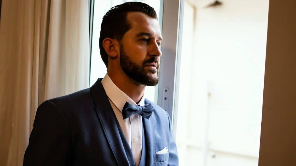 A serious, well-dressed man in a blue suit and bow tie looks out a window.