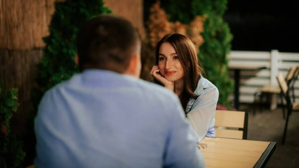 A smiling woman looks intently at a man sitting across from her at a table outdoors at night.